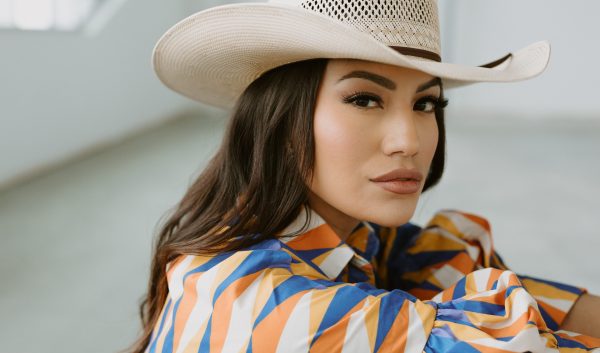 Ashley Callingbull smolders at the camera, wearing a cream cowgirl hat and dress with sharp geometric patterns of cream, orange, yellow, and deep blue.